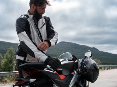 HALVARSSONS SOLBERG Person with beard standing over a motorcycle. White jacket on show. Mountains in background.